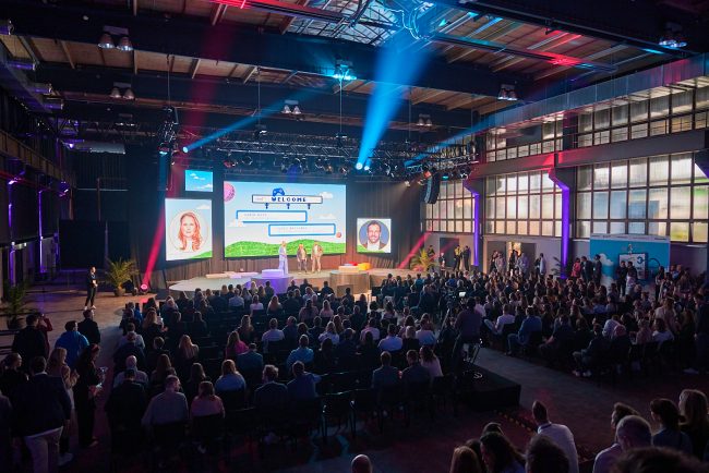 Eventlocation, die voll mit Menschen ist und vorne Speaker einen Vortrag halten